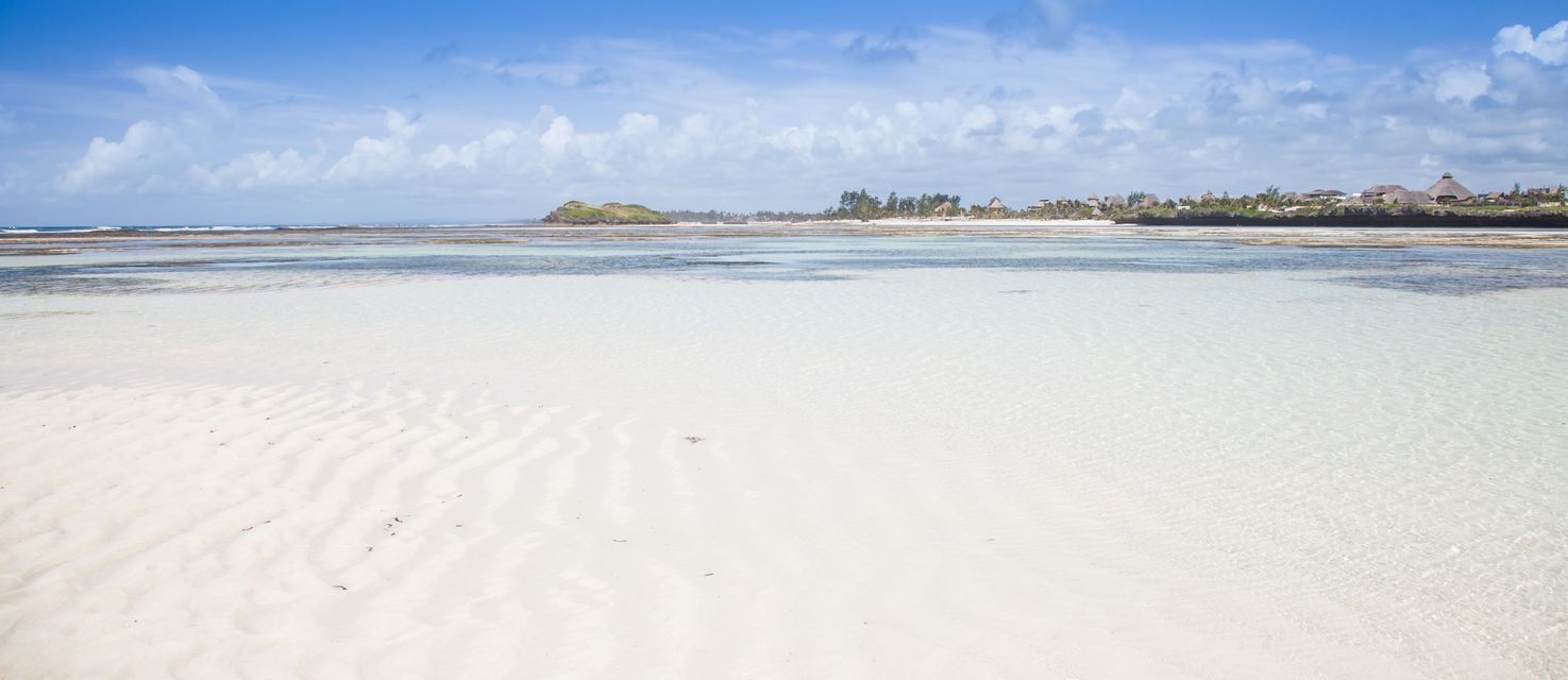 Una amplia extensión de arena blanca con ondulaciones es visible bajo aguas claras y poco profundas, con una costa tropical a lo lejos bajo un cielo azul.
