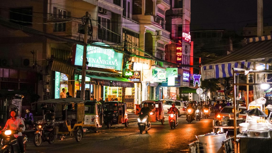 Eine belebte Stadtstraße bei Nacht mit Verkehr von Motorrädern und Tuk-Tuks unter bunten Leuchtreklamen.