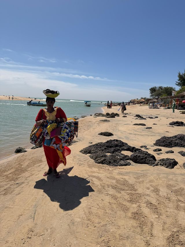 Una mujer con un vestido rojo camina por una playa de arena, equilibrando una cesta en la cabeza y llevando coloridas bolsas de tela.
