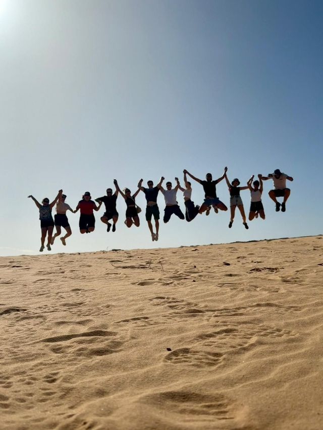 Un viaje en grupo de WeRoad, cogidos de la mano y saltando juntos en el aire en una duna de arena bajo un cielo brillante y despejado.