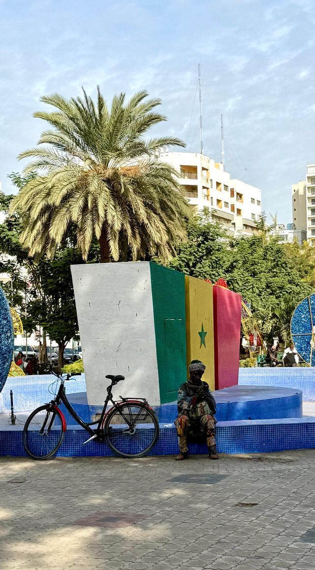 Una persona con ropa estampada se sienta en el borde de una base de azulejos azules junto a un gran monumento pintado con la bandera senegalesa.