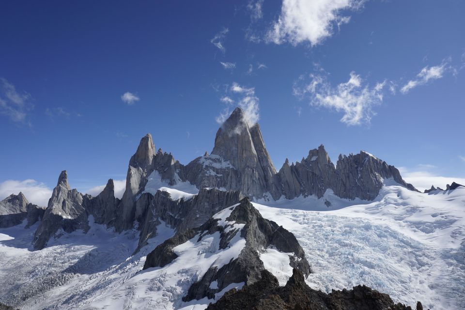A range of jagged, rocky mountains covered in snow and glaciers under a bright blue sky with sparse clouds.