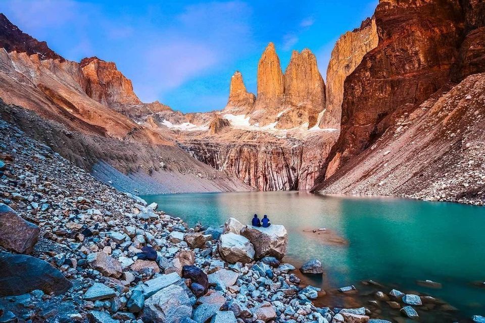 Due persone siedono su un grande masso sulla riva di un lago turchese, ai piedi di drammatiche guglie montane illuminate dal sole.