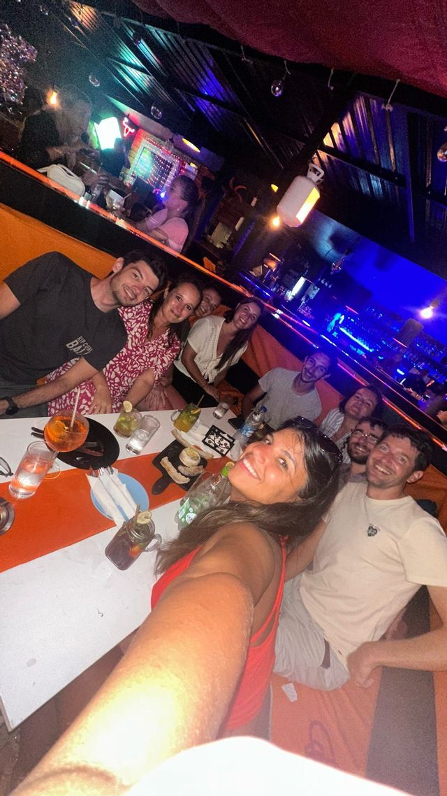 Un groupe WeRoad prend un selfie, assis à une longue table avec des boissons, dans une boîte de nuit à l'ambiance tamisée.