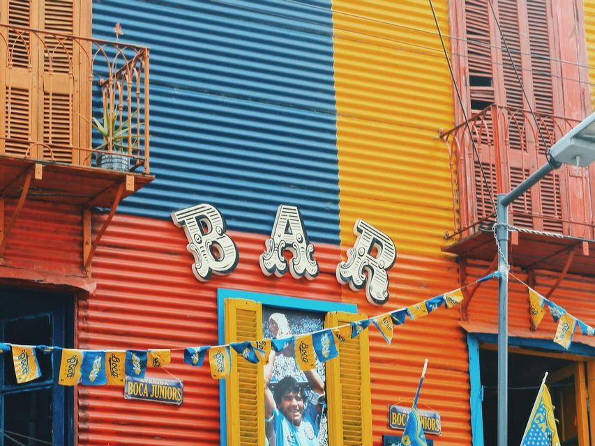 Una colorida fachada de edificio de metal corrugado en rojo, azul y amarillo, con un letrero de 'BAR' y pequeñas banderas colgadas.