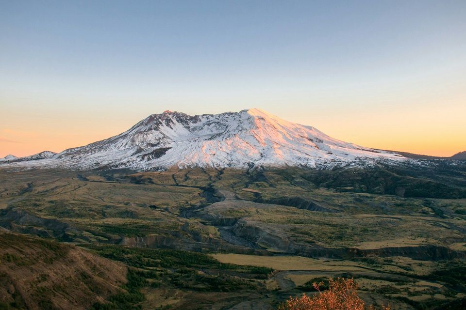 Una cima montuosa innevata è illuminata dall'alba, con una vasta valle che si estende sotto un cielo sereno.