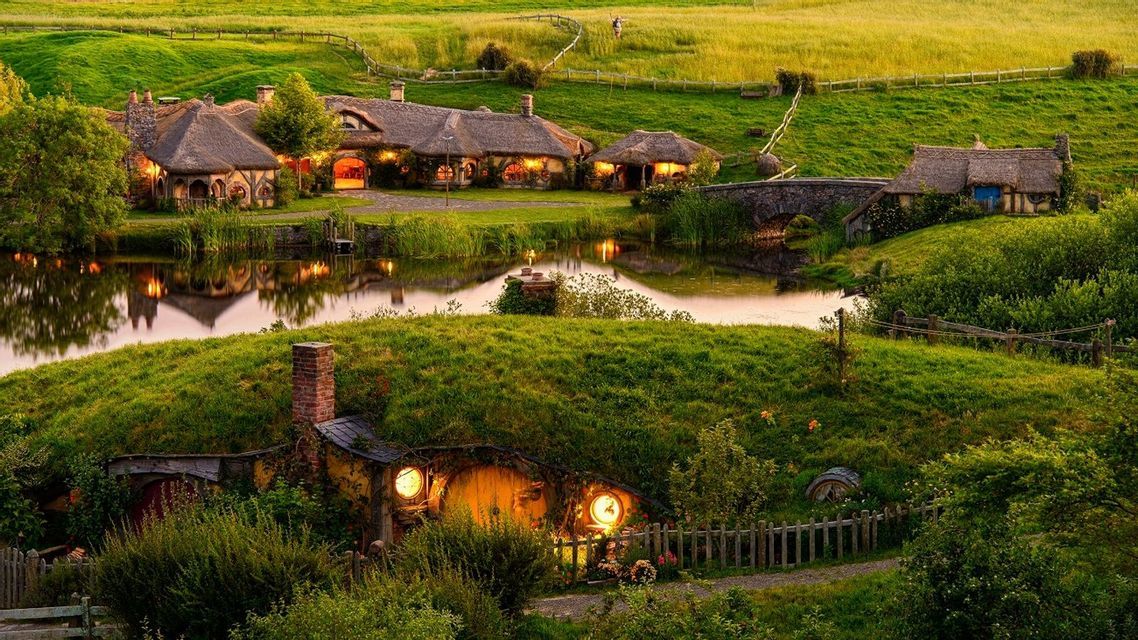 A village of houses with thatched roofs and glowing windows is nestled into lush green hillsides beside a still pond.
