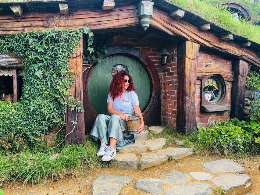 A woman with curly red hair sits on the stone steps in front of a house with a round green door and a grass-covered roof.
