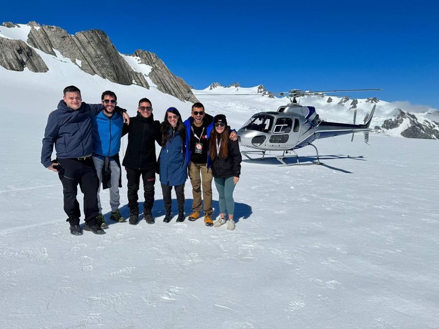 Un groupe WeRoad souriant sur un glacier enneigé, avec un hélicoptère et des montagnes en arrière-plan.