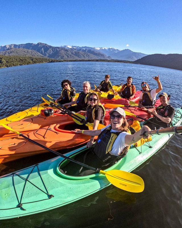 Un voyage en groupe WeRoad prend un selfie en faisant du kayak sur un lac calme avec des montagnes en arrière-plan.