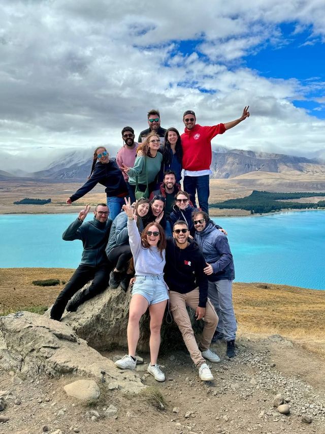 Un groupe WeRoad en voyage posant pour une photo sur une colline rocheuse, avec un lac turquoise et des montagnes en arrière-plan sous un ciel nuageux.