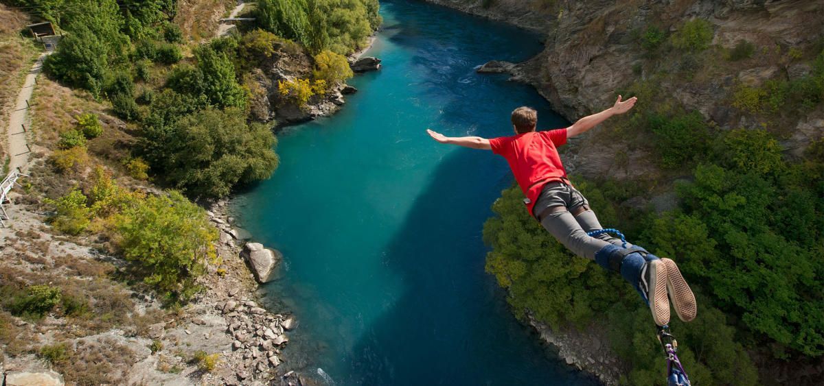 Eine Person im roten Hemd beim Bungee-Jumping mit ausgestreckten Armen über einem türkisfarbenen Fluss in einer felsigen Schlucht.