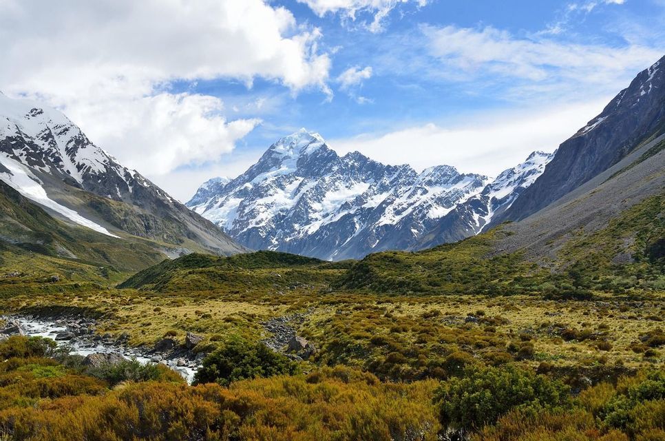 A snow-capped mountain range rises above a vast valley filled with green and gold shrubs, with a small stream flowing through it.