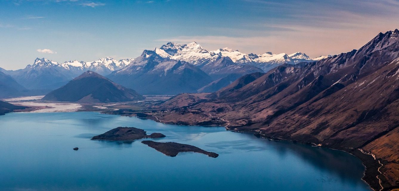 An aerial view of a large blue lake with small islands, surrounded by rugged mountains with snow-capped peaks in the background.