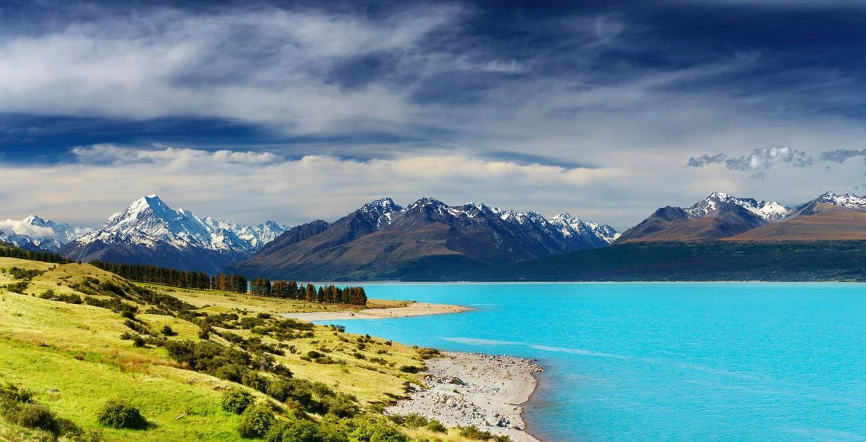 A grassy shore meets a vibrant turquoise lake in front of a distant, snow-capped mountain range under a partly cloudy sky.