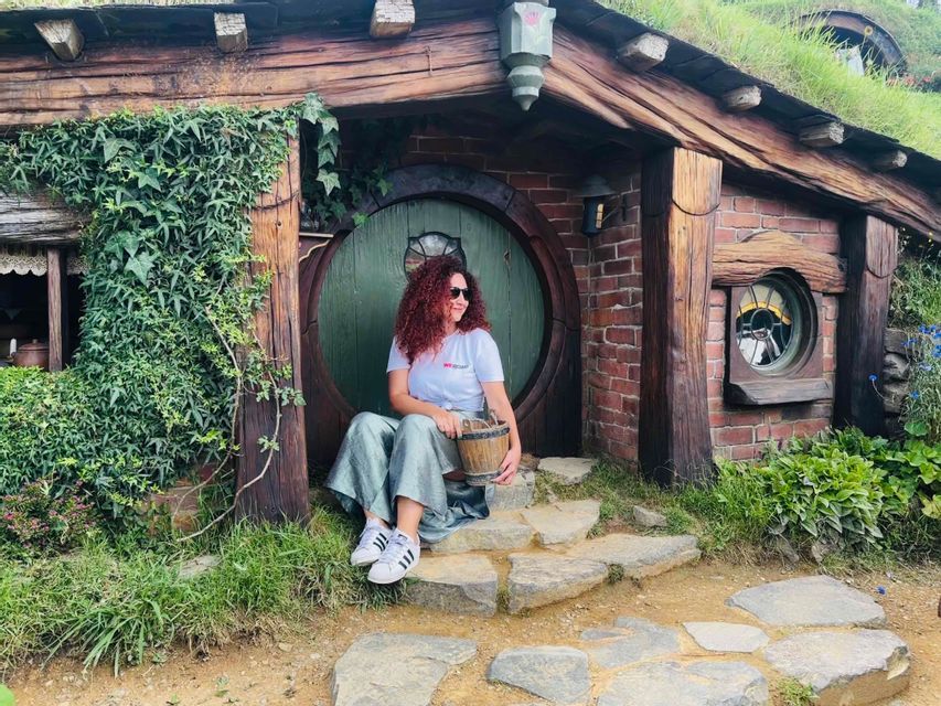 Une femme aux cheveux roux bouclés est assise sur les marches en pierre d'une maison de style hobbit, tenant un petit seau en bois.