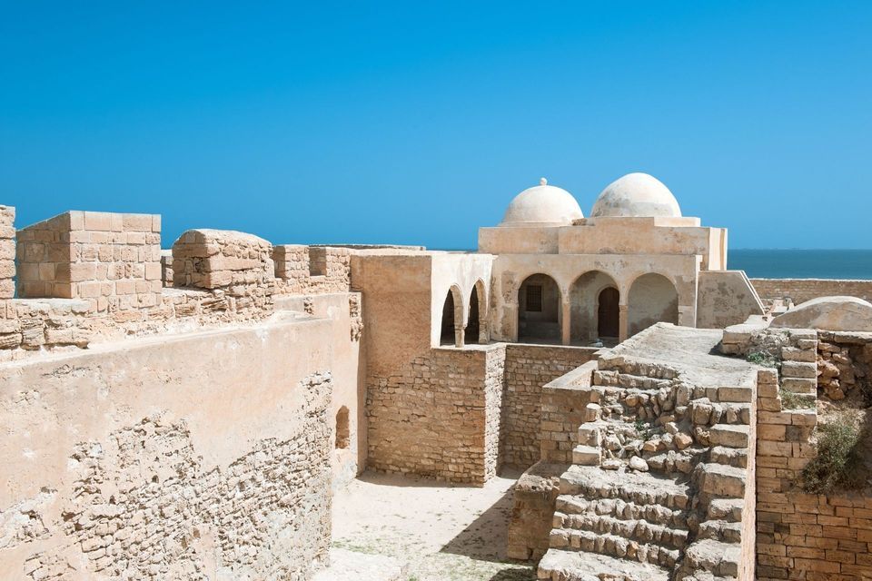Antike, sandfarbene Steinruinen einer Küstenfestung mit Kuppelbauten unter klarem blauen Himmel.
