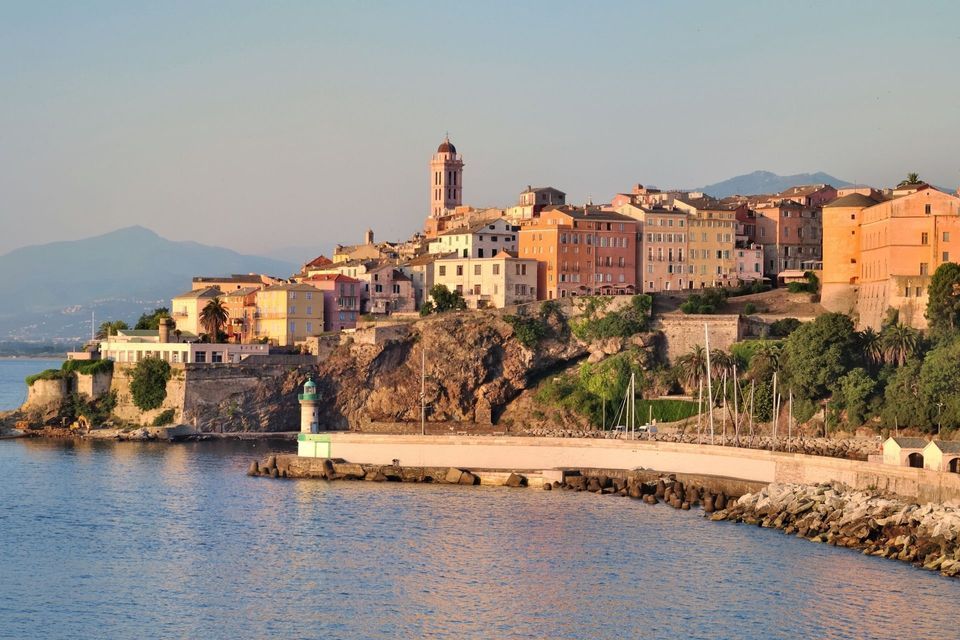 Eine Küstenstadt mit farbenfrohen Gebäuden und einem Glockenturm liegt auf einer felsigen Klippe mit Blick auf das Meer und einem kleinen Leuchtturm auf einem Pier.