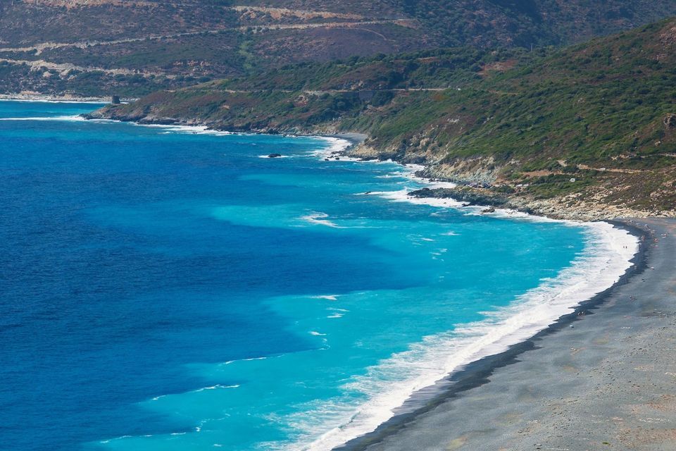 Eine Luftaufnahme einer Küstenlinie, wo lebhaft türkisfarbenes und tiefblaues Wasser auf einen dunklen Kiesstrand trifft.