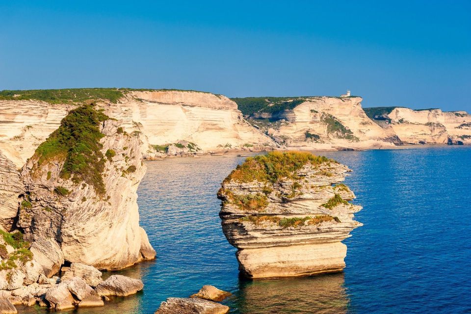 Weiße Kalksteinklippen und eine große, grün bewachsene Felsnadel im Meer, vor einem ruhigen blauen Meer unter klarem Himmel.