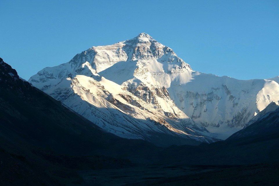 Una grande cima montuosa coperta di neve è illuminata dal sole contro un cielo azzurro limpido, con una valle scura e in ombra in primo piano.