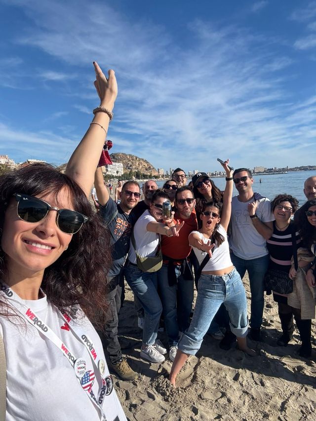 Un viaggio di gruppo WeRoad scatta un selfie di gruppo su una spiaggia sabbiosa, con una città costiera e il mare alle loro spalle.