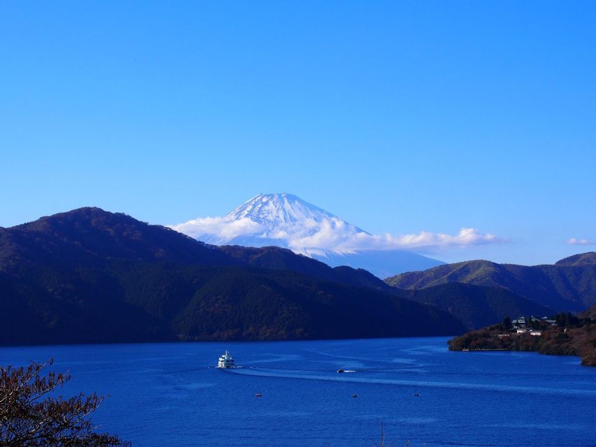 Un traghetto bianco naviga su un lago azzurro, con montagne boscose e il Monte Fuji innevato visibili sullo sfondo.