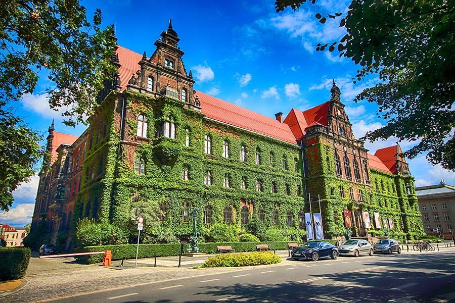 Un bâtiment en briques orné, avec un toit rouge et sa façade couverte de lierre vert, se dresse à côté d'une rue sous un ciel bleu éclatant.