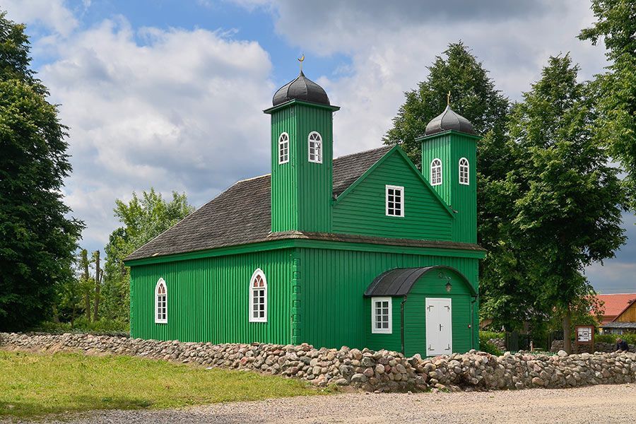 Une mosquée en bois verte à deux minarets se dresse derrière un bas mur de pierre, entourée d'arbres sous un ciel partiellement nuageux.