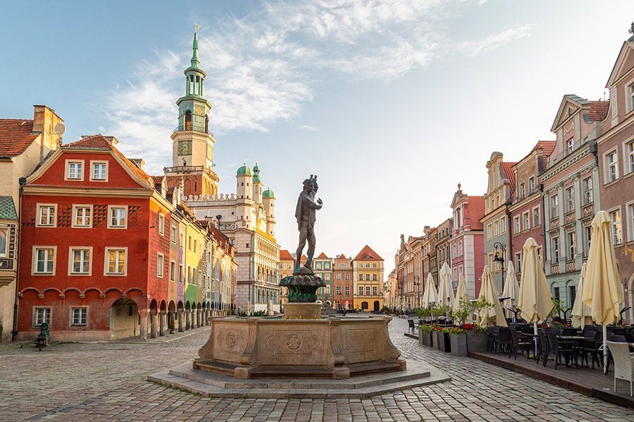 Une fontaine ornée avec une statue au milieu d'une place historique pavée, entourée de bâtiments colorés et d'une tour de l'horloge au lever du soleil.