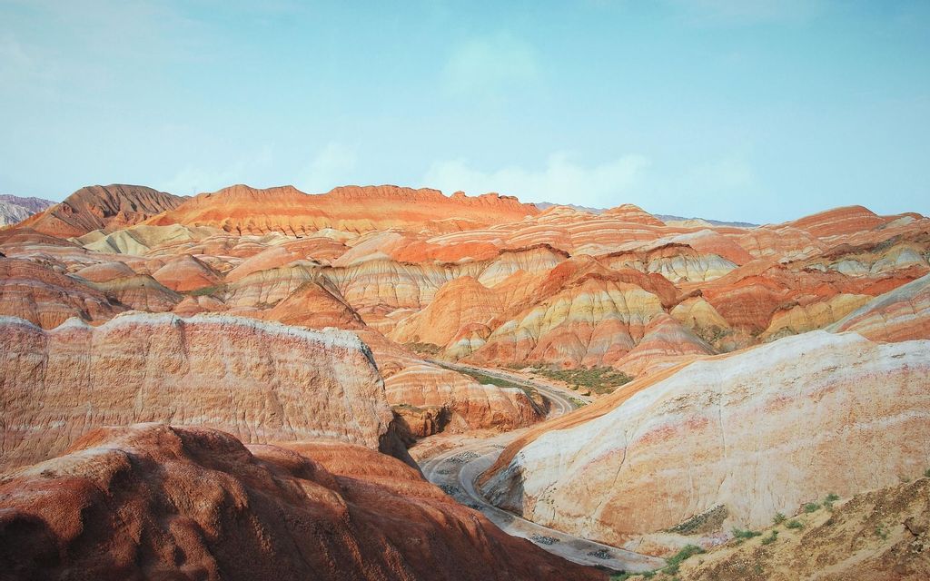 Un vasto paesaggio di montagne colorate e stratificate con sfumature di arancione, rosso e giallo sotto un cielo azzurro chiaro.