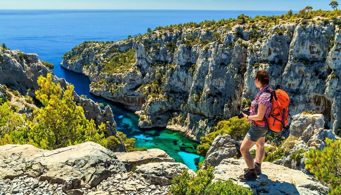 Una donna con uno zaino arancione si trova su una scogliera rocciosa, ammirando un'insenatura di acqua turchese circondata da scogliere.