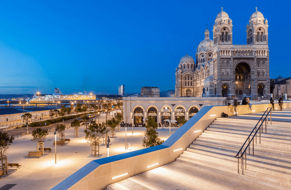 Una cattedrale illuminata con pietra striata si affaccia su una moderna piazza e un porto con una nave da crociera al tramonto.
