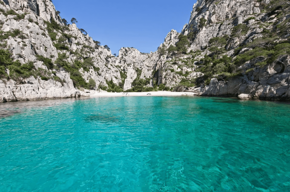 Acqua turchese in una caletta appartata con una piccola spiaggia sabbiosa, circondata da ripide scogliere rocciose sotto un cielo azzurro e limpido.