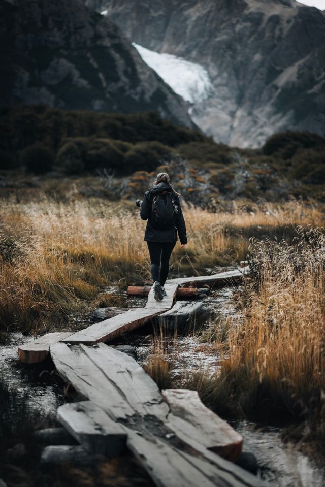 Un escursionista con uno zaino cammina su una passerella di legno attraverso un campo, con montagne e un ghiacciaio sullo sfondo.