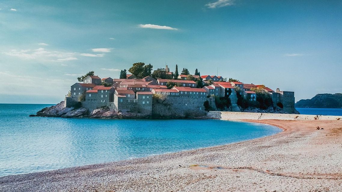 Un borgo isolano fortificato con edifici in pietra e tetti di tegole rosse, ammirato da una spiaggia di ciottoli su un mare calmo e turchese.
