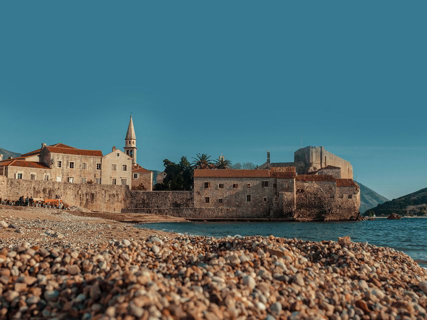 Veduta di una storica città in pietra con una fortezza sulla costa, da una spiaggia di ciottoli sotto un cielo azzurro e limpido.
