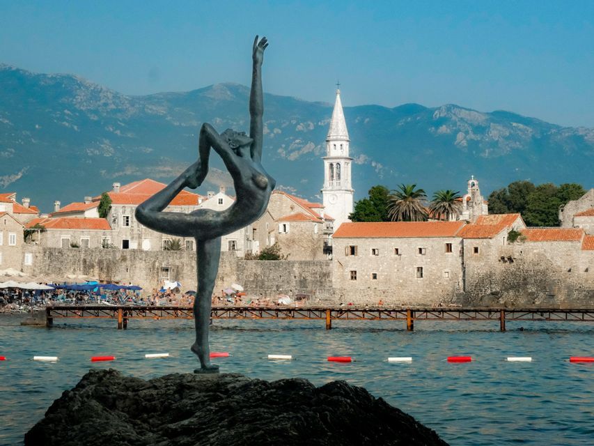 Una statua di bronzo di una ballerina in posa su una roccia nell'acqua, con una storica città costiera e montagne sullo sfondo.