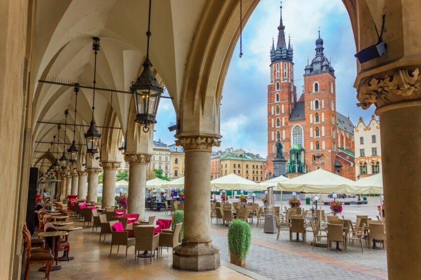 Vue d'une terrasse de café installée sous une colonnade de pierre voûtée, sur une place de ville historique, avec une grande église en briques en arrière-plan.