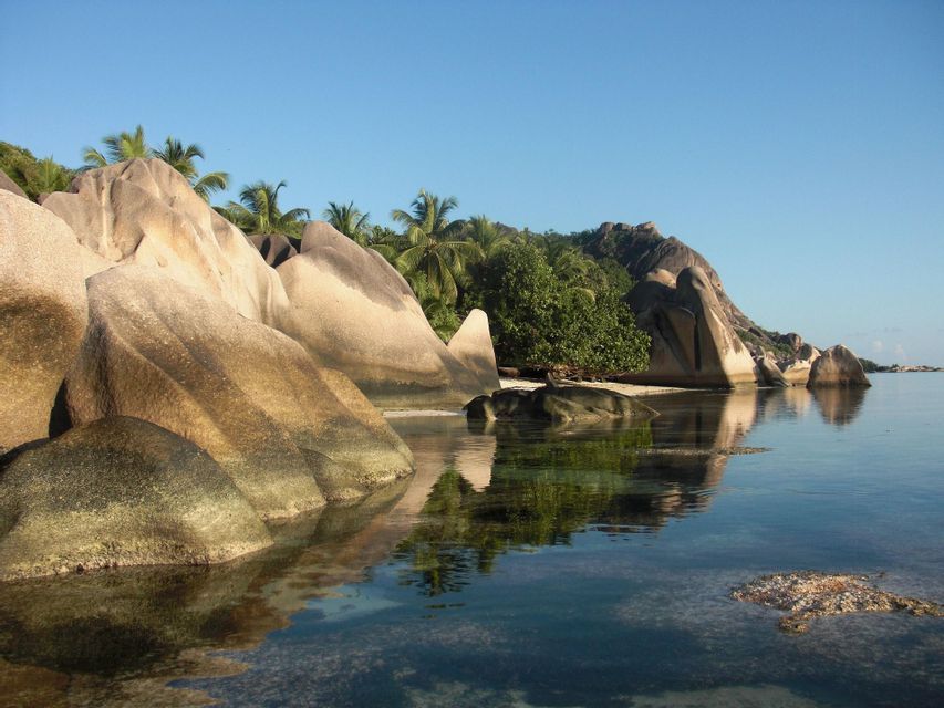 Große, glatte Granitfelsen liegen an einer tropischen Küste und spiegeln sich im ruhigen, klaren Wasser unter blauem Himmel wider.