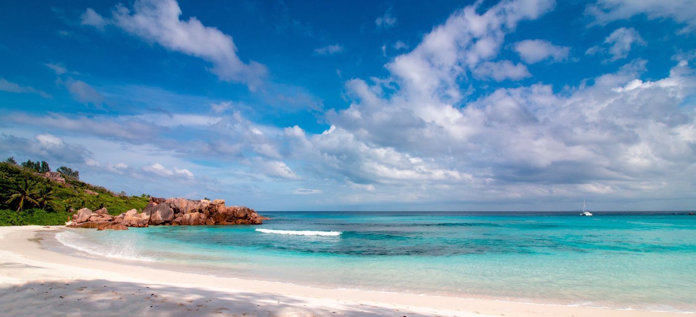 Ein tropischer Strand mit weißem Sand und türkisfarbenem Wasser schmiegt sich an eine felsige Küste mit Palmen unter einem blauen, bewölkten Himmel.
