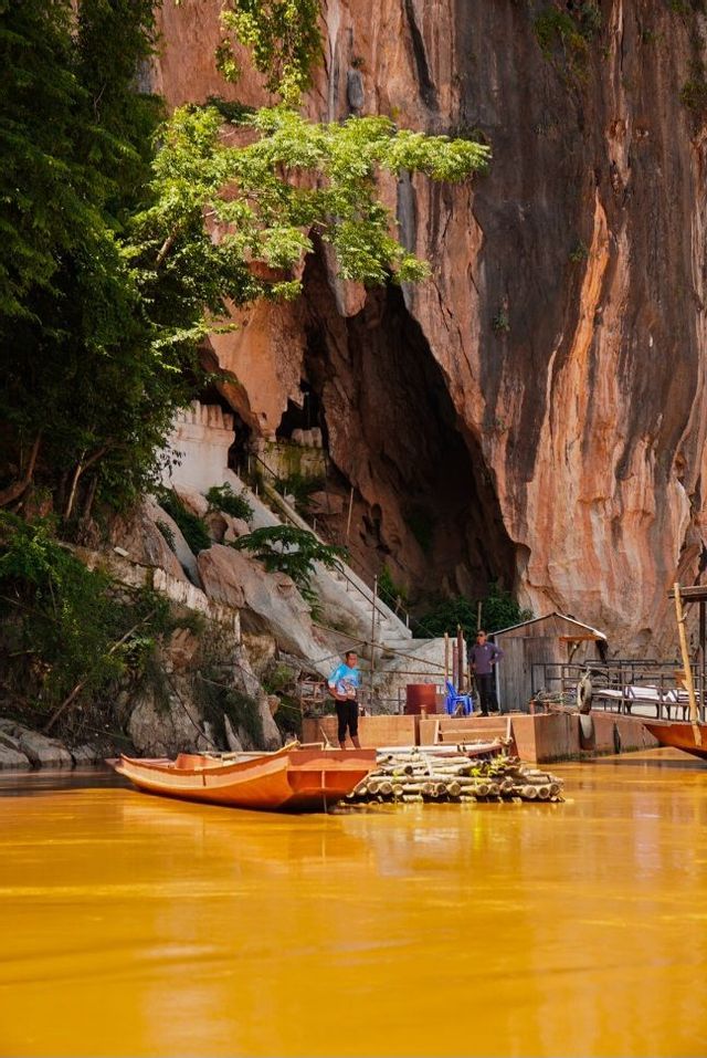 Un bateau en bois est amarré sur une rivière jaune au pied d'une grande falaise avec une entrée de grotte et un petit bâtiment.