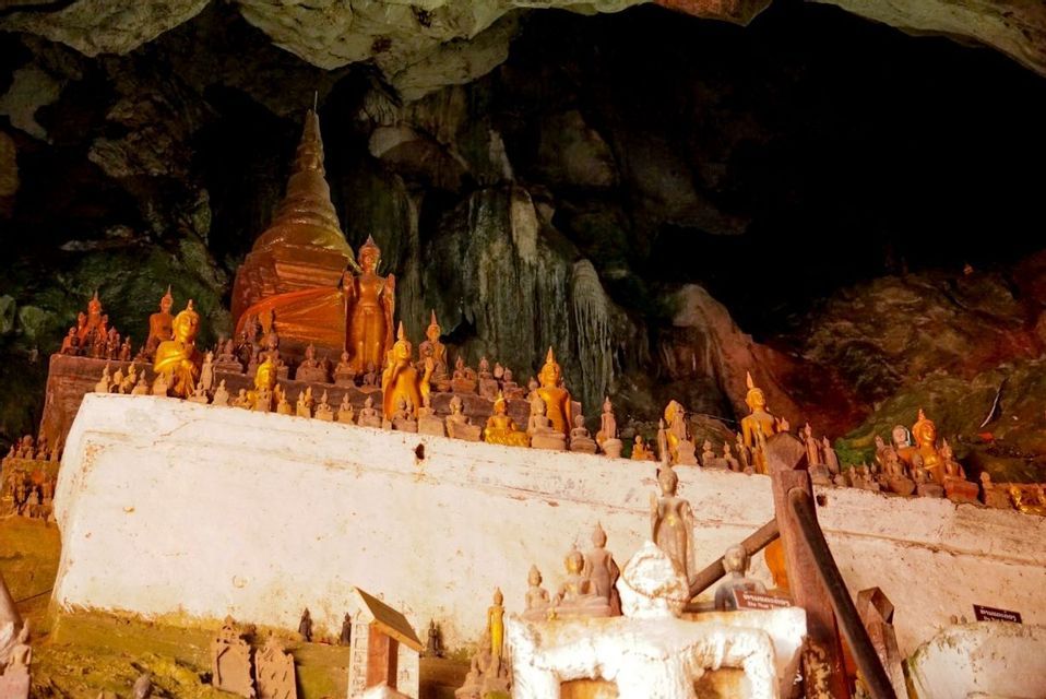 De nombreuses statues de Bouddha dorées et un stupa exposés sur une plateforme blanche à l'intérieur d'une grotte sombre et rocheuse.