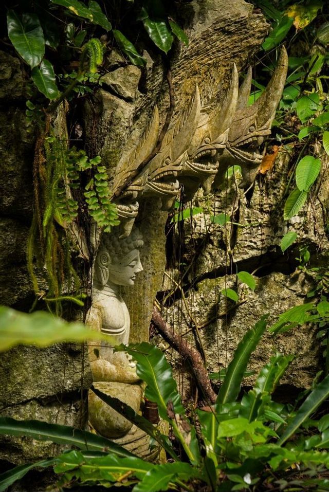 Une statue de Bouddha en pierre abritée par des têtes de Naga sculptées, nichée dans une alcôve rocheuse entourée d'un feuillage luxuriant de la jungle.