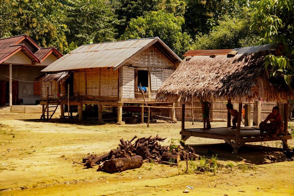 Des maisons en bois sur pilotis et une hutte au toit de chaume se dressent dans une clairière poussiéreuse d'un village, avec une forêt dense en arrière-plan.