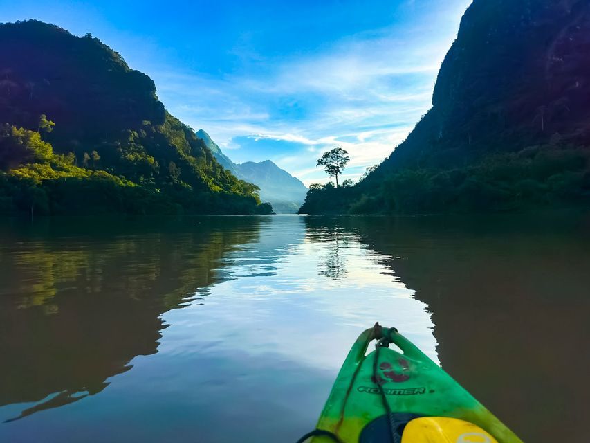 Vue à la première personne depuis un kayak vert sur une rivière calme, bordée de montagnes escarpées et boisées, sous un ciel bleu éclatant.
