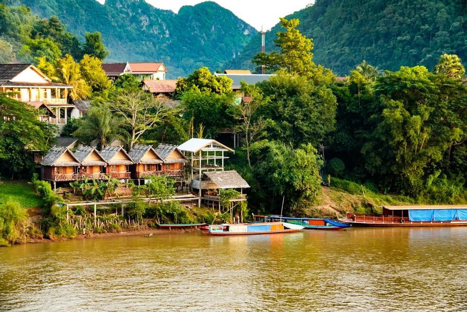 Un village de bungalows en bois sur pilotis est installé au bord d'une rivière, entouré d'une jungle luxuriante et de montagnes, avec plusieurs bateaux amarrés sur l'eau.