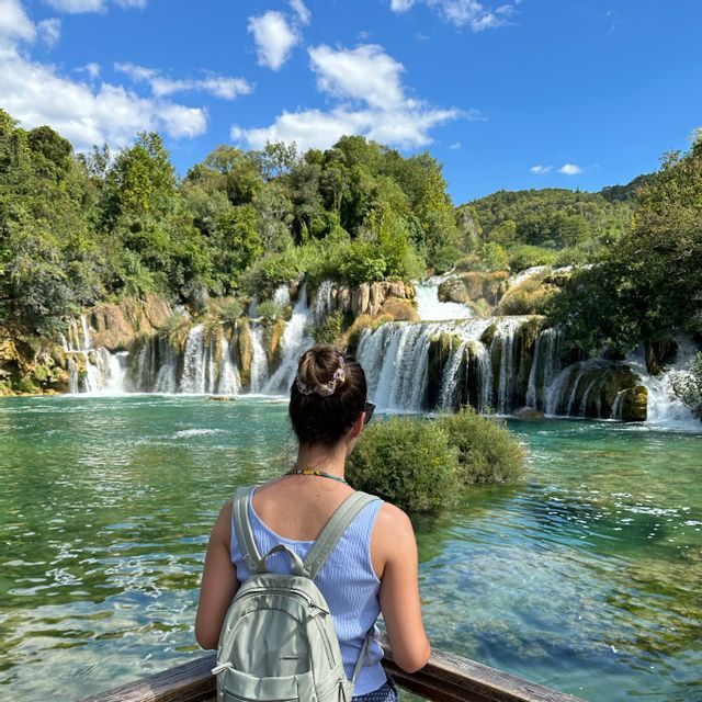 Eine Frau mit Rucksack, von hinten gesehen, blickt auf einen weiten, kaskadierenden Wasserfall, umgeben von üppigen grünen Bäumen.