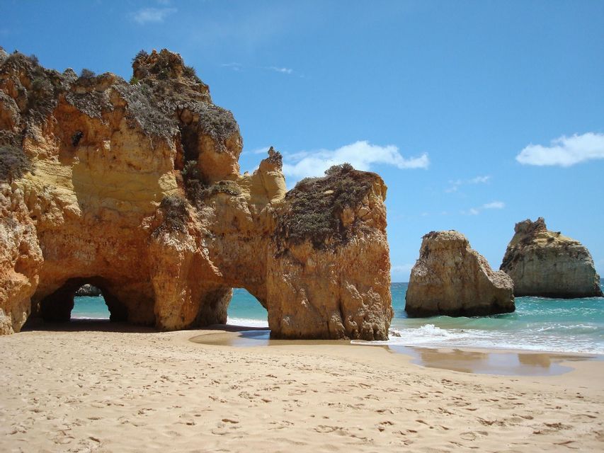 Grandi formazioni rocciose arancioni con archi naturali su una spiaggia sabbiosa, con acqua turchese e un cielo blu sullo sfondo.