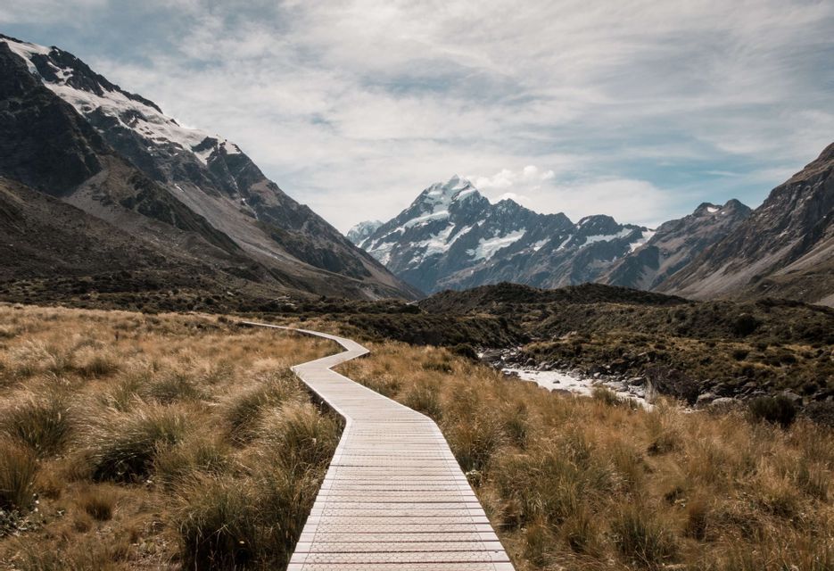 Une promenade en bois sinueuse mène à travers une vallée herbeuse vers de hautes montagnes enneigées sous un ciel nuageux.